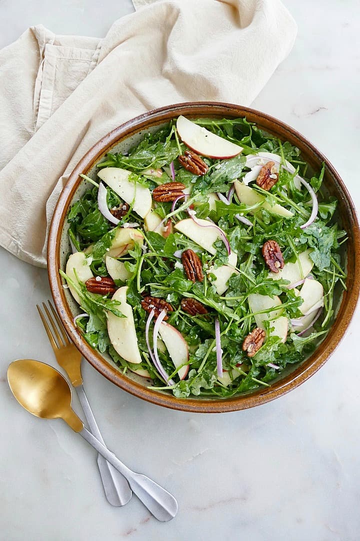 arugula apple salad in a large serving bowl next to gold and silver utensils