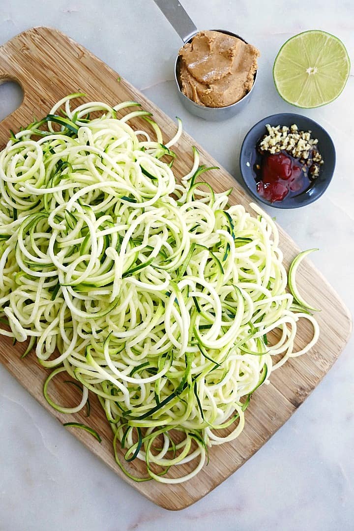zucchini noodles on a bamboo cutting board next to a lime, peanut butter, and other seasonings