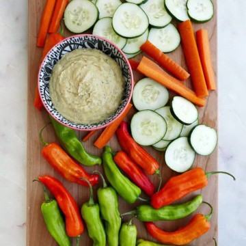 a bowl of dip, sliced carrots, cucumbers, and peppers on a serving tray