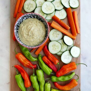 a bowl of dip, sliced carrots, cucumbers, and peppers on a serving tray