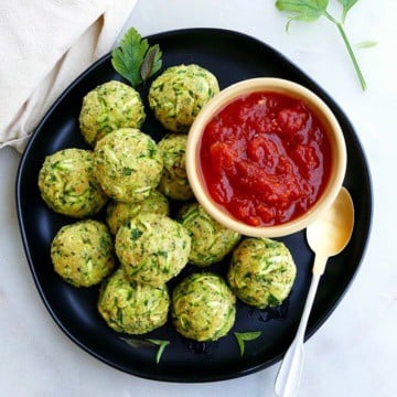 zucchini balls on a serving plate with tomato sauce in a small bowl