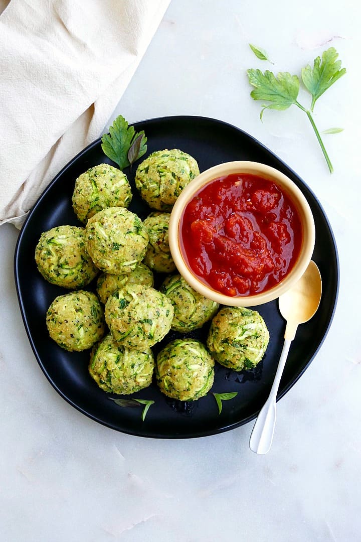 zucchini balls on a serving plate with tomato sauce in a small bowl