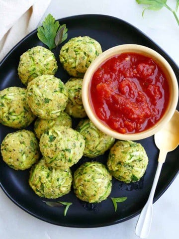 square image of zucchini balls on a serving plate with a bowl of tomato sauce