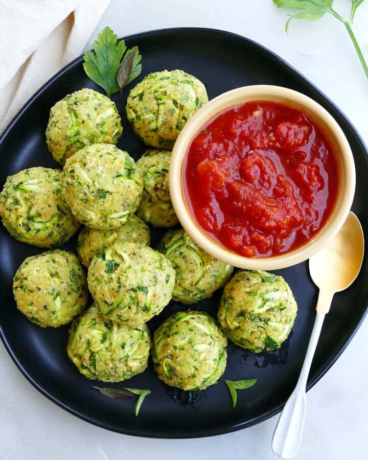 square image of zucchini balls on a serving plate with a bowl of tomato sauce