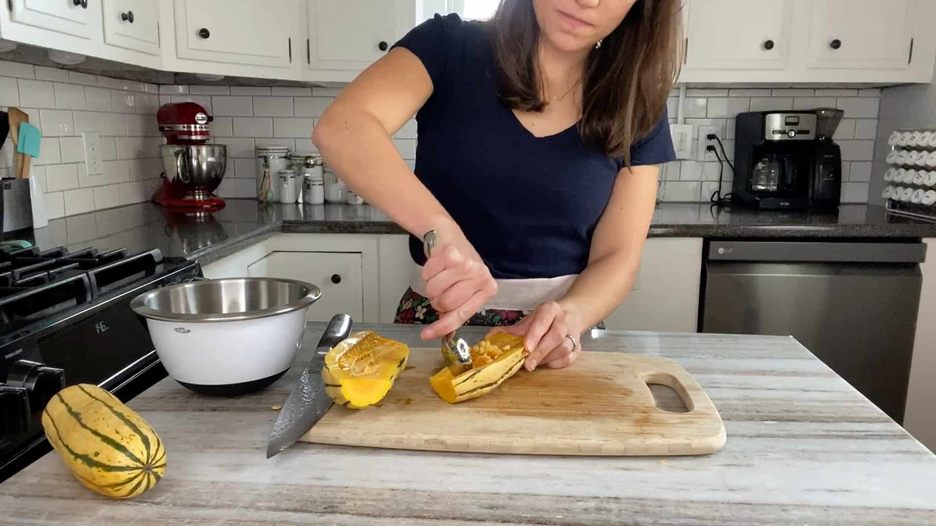 woman scooping out seeds from half of a delicata squash over a cutting board