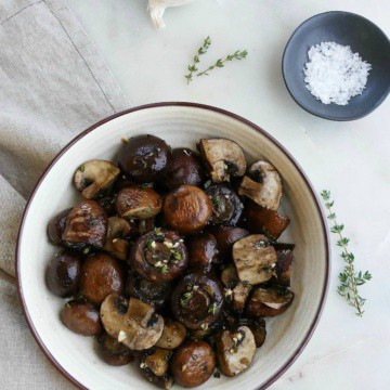 serving bowl with garlic roasted mushrooms next to garlic bulbs, thyme, and salt