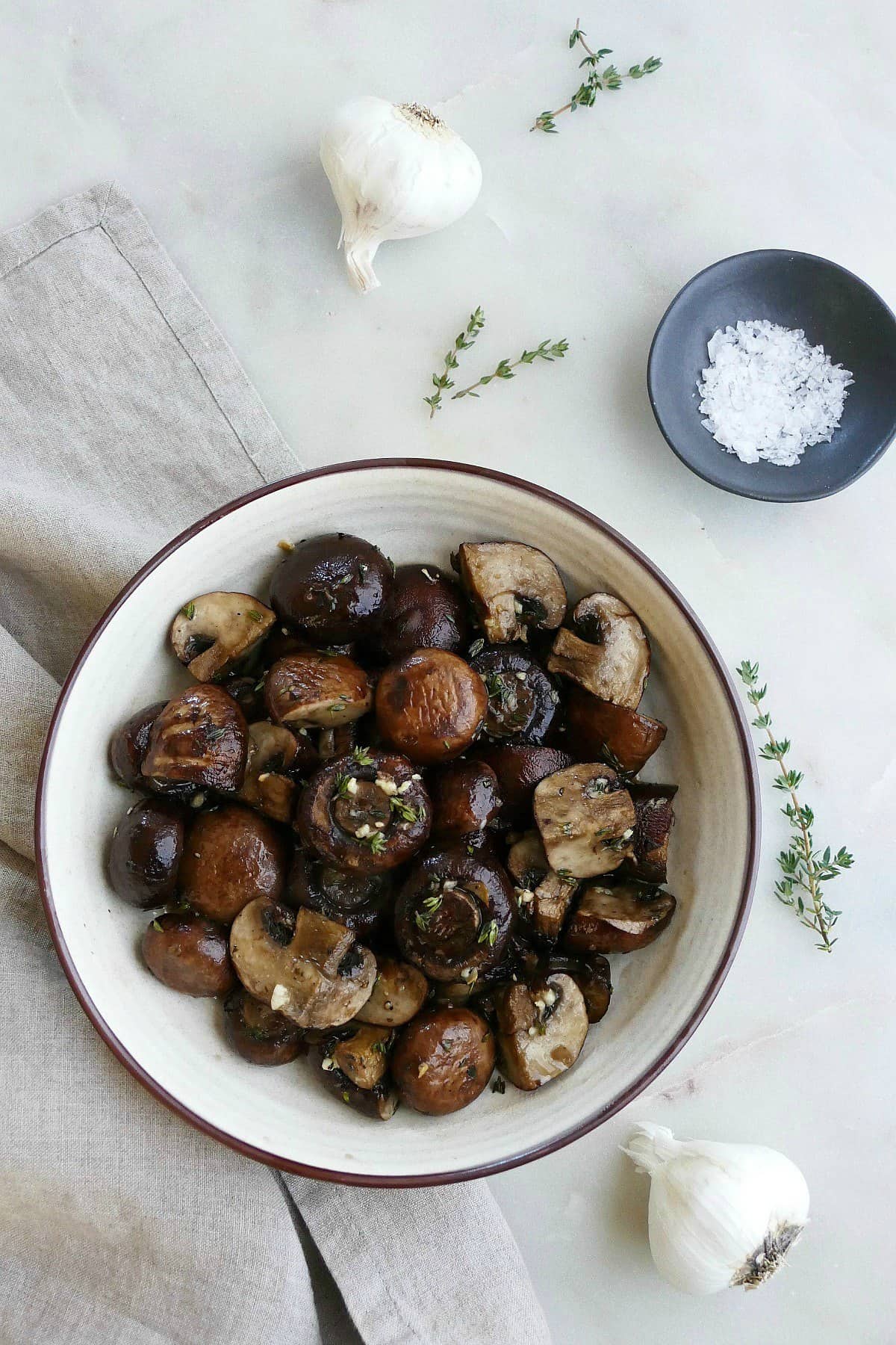 serving bowl with garlic roasted mushrooms next to garlic bulbs, thyme, and salt