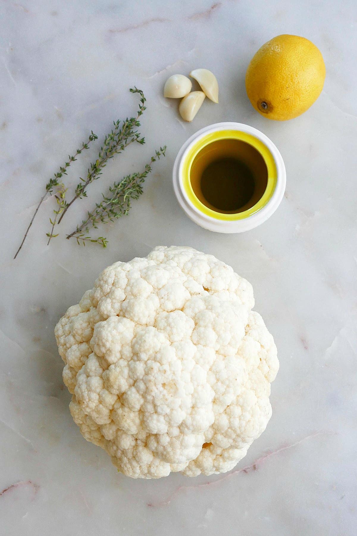 head of cauliflower next to herbs, garlic, lemon, and oil on a counter