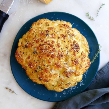 baked whole cauliflower on a serving plate next to parmesan cheese and napkin
