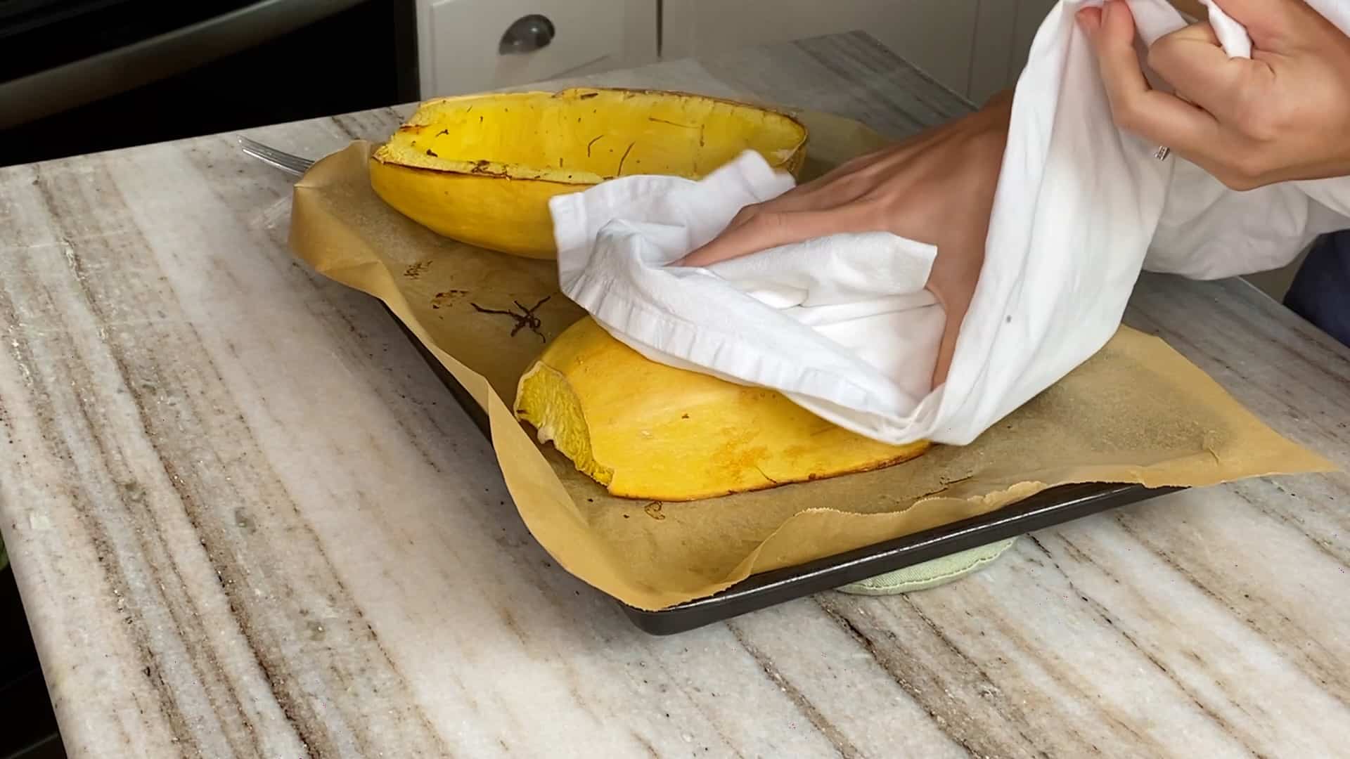 woman flipping spaghetti squash over with a dish towel over a baking sheet