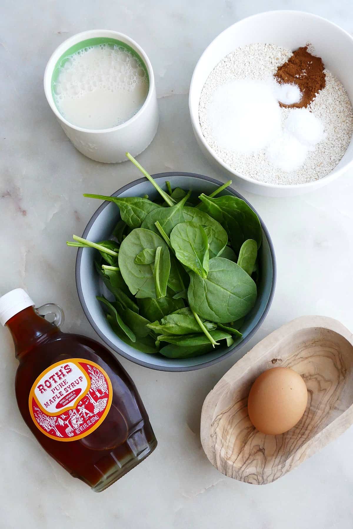 spinach, milk, dry ingredients, and an egg in bowls next to a bottle of syrup