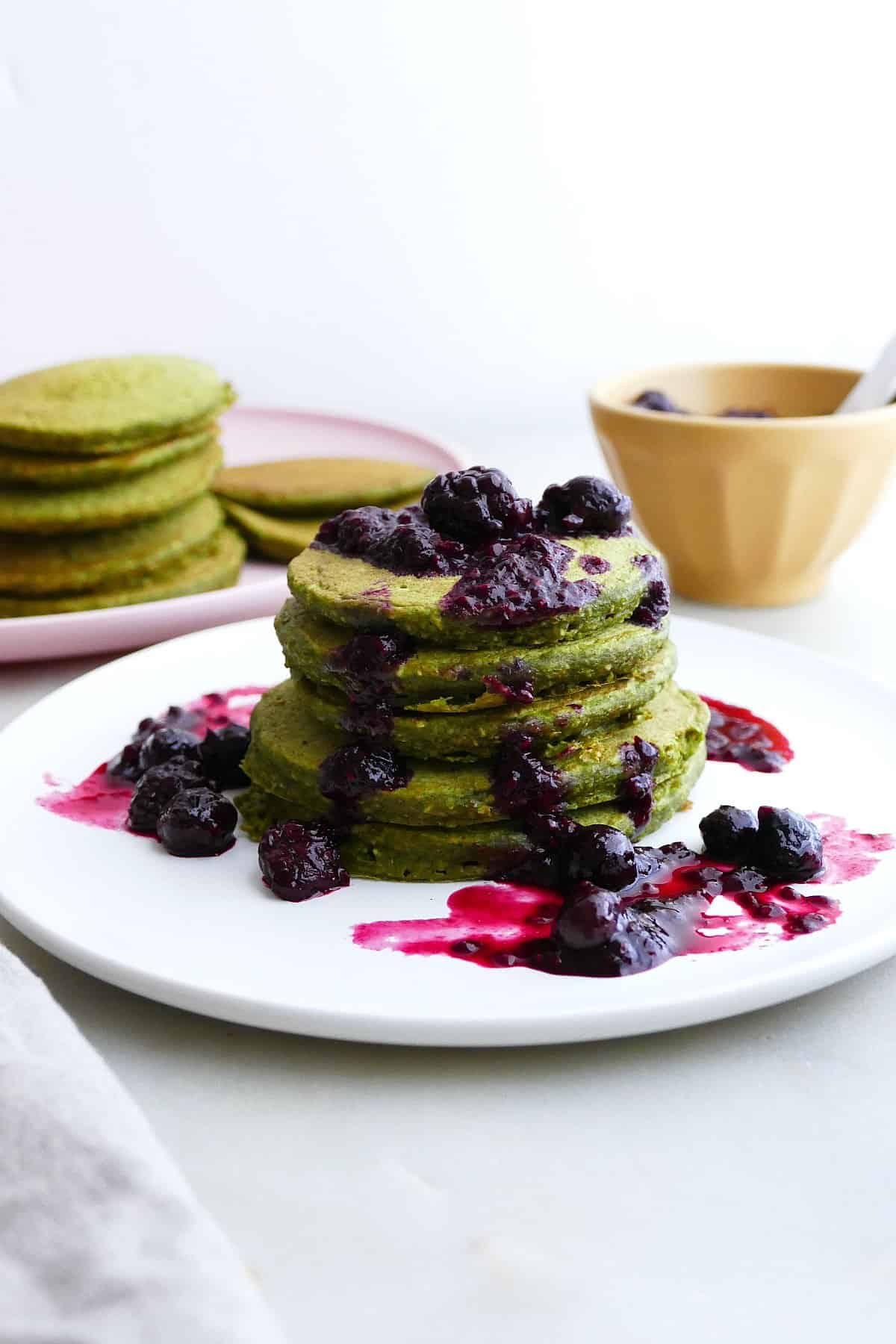 stack of spinach pancakes with berry compote on a plate in front of more pancakes