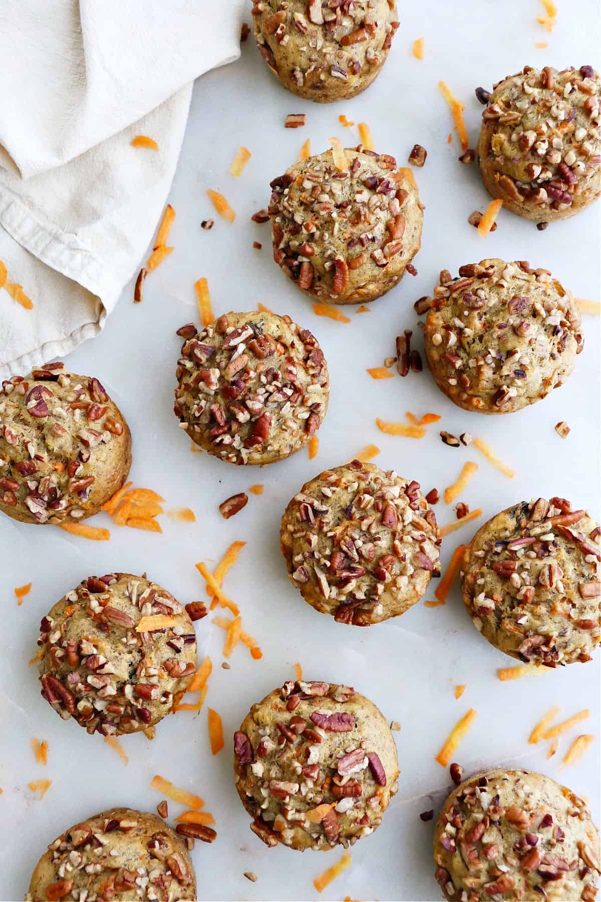 12 banana carrot muffins spread out on a counter with pecans and shredded carrots