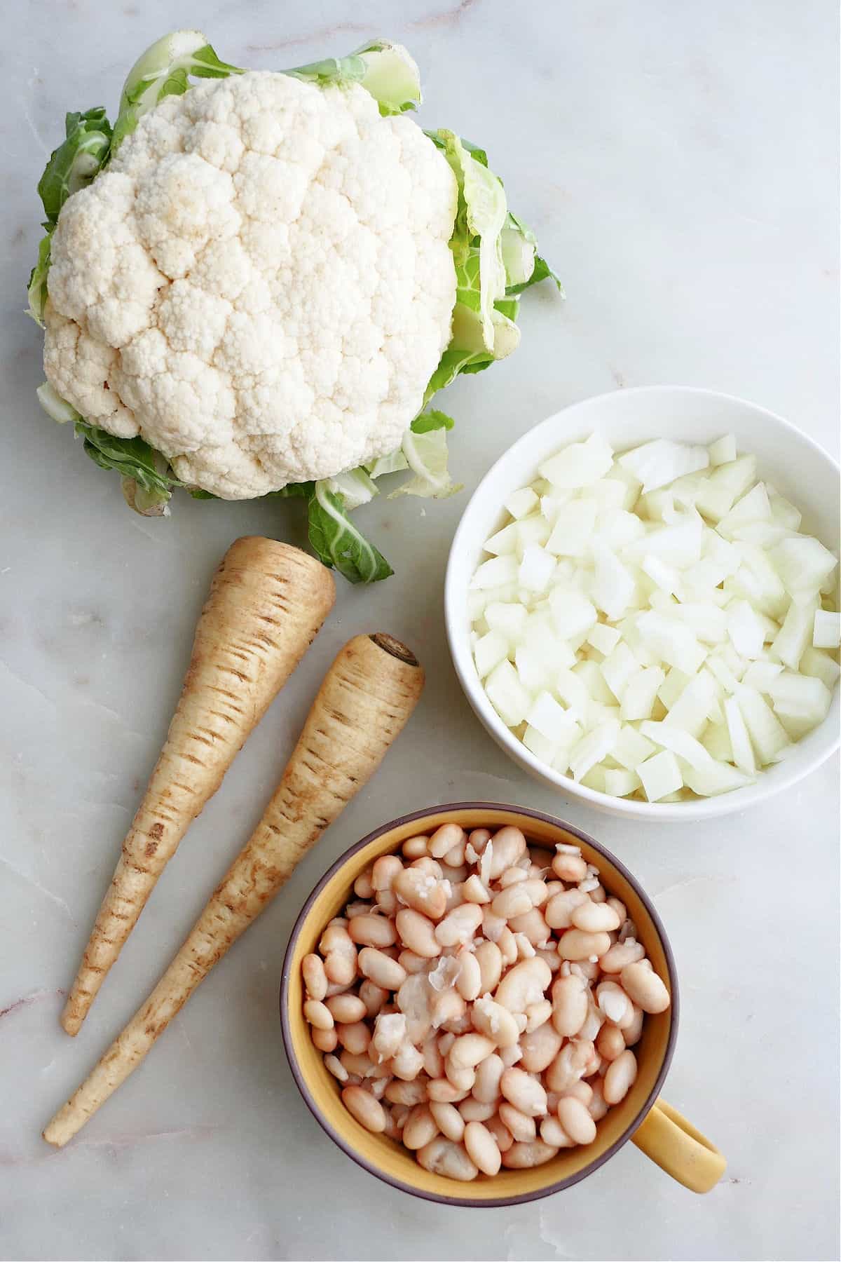 cauliflower, two parsnips, diced onion, and white beans next to each other on a counter