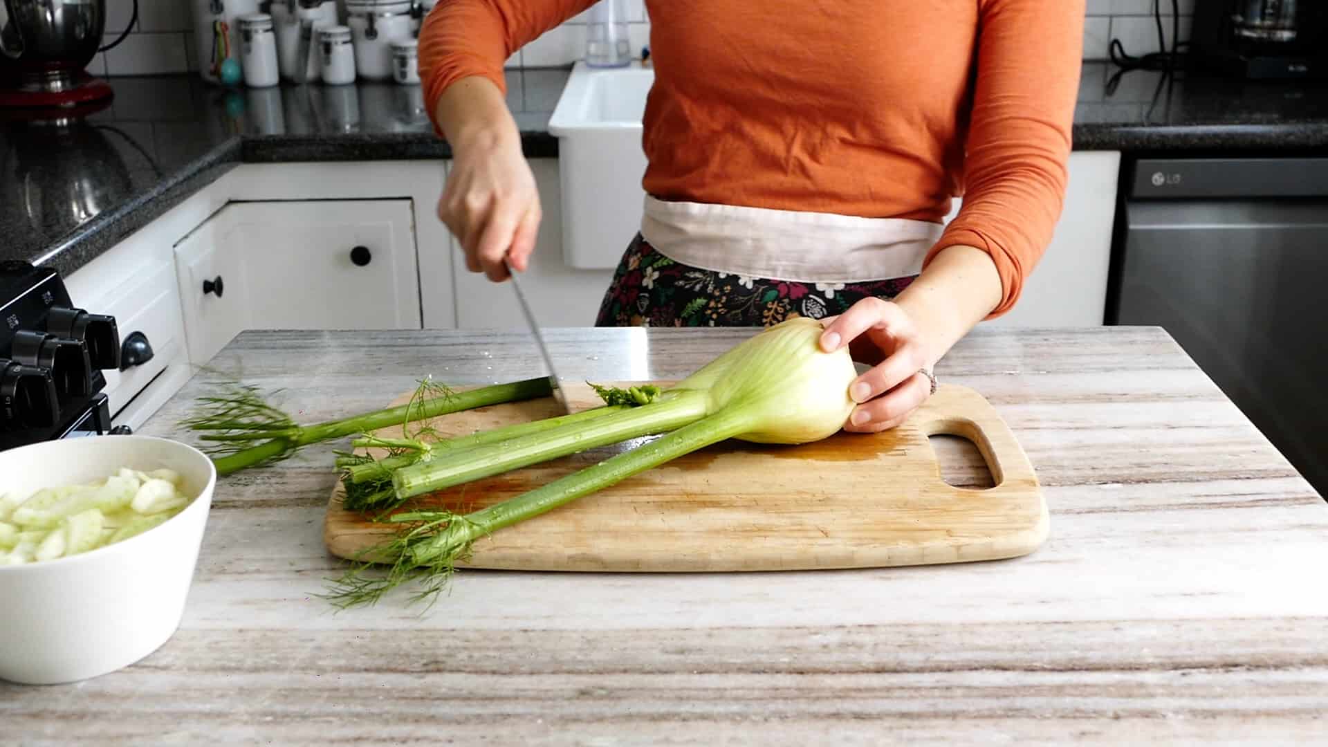 woman cutting stalks and fronds off of a bulb of fennel in a kitchen