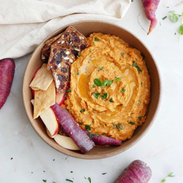 carrot hummus in a serving bowl with crackers, apples, and sliced carrots on a counter