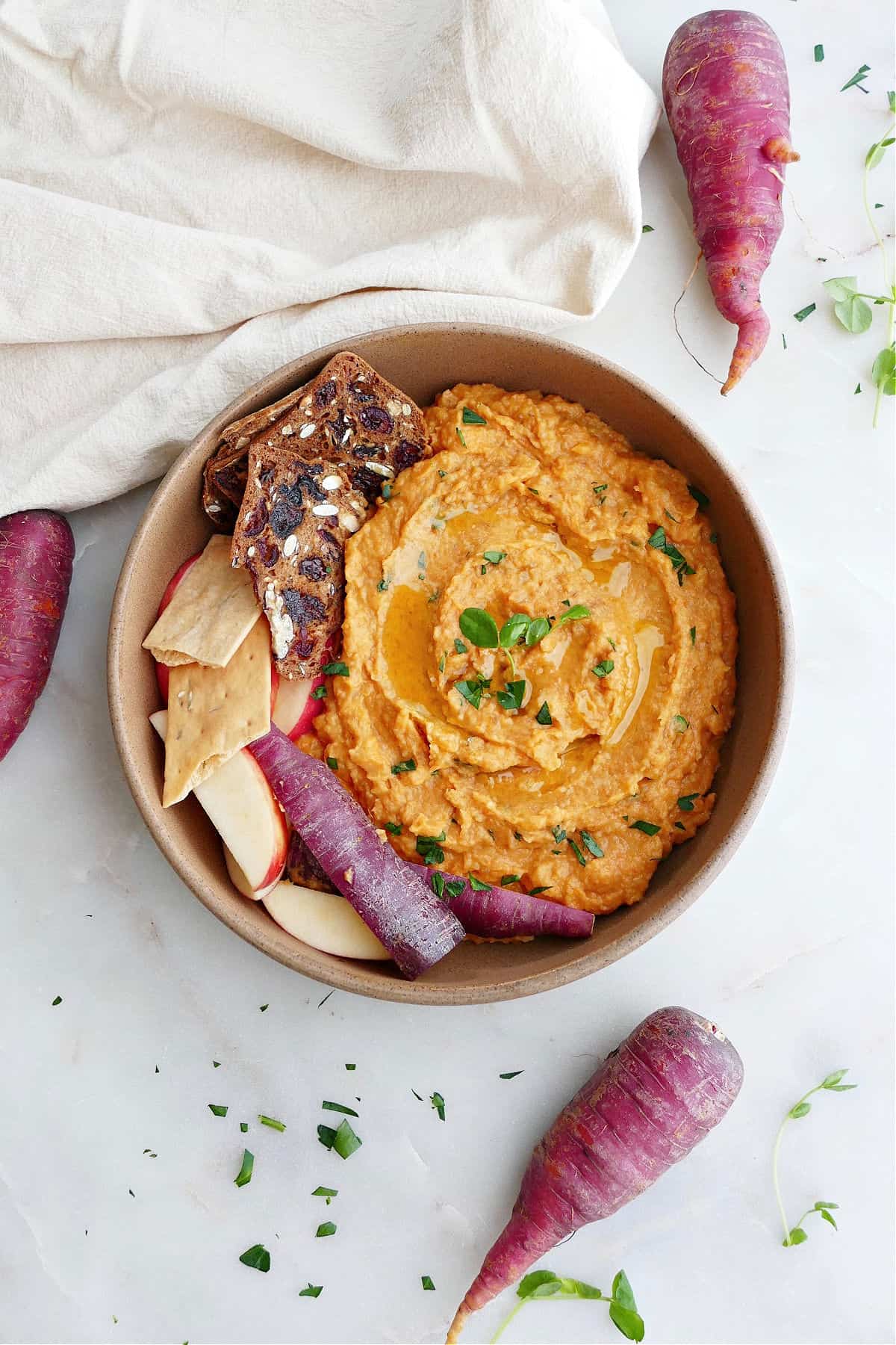 carrot hummus in a serving bowl with crackers, apples, and sliced carrots on a counter