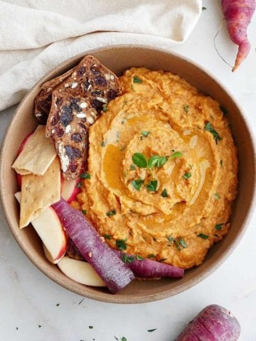 square image of purple carrot hummus in a serving bowl with crackers, apples, and sliced carrots