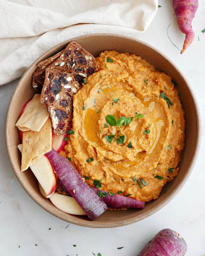 square image of purple carrot hummus in a serving bowl with crackers, apples, and sliced carrots