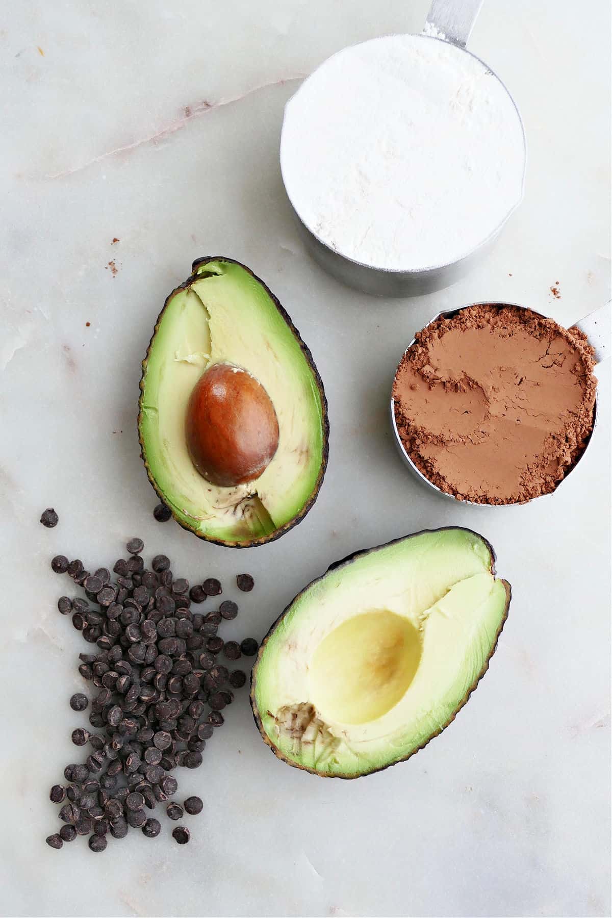 two avocado halves, chocolate chips, flour, and cocoa powder next to each other on a counter