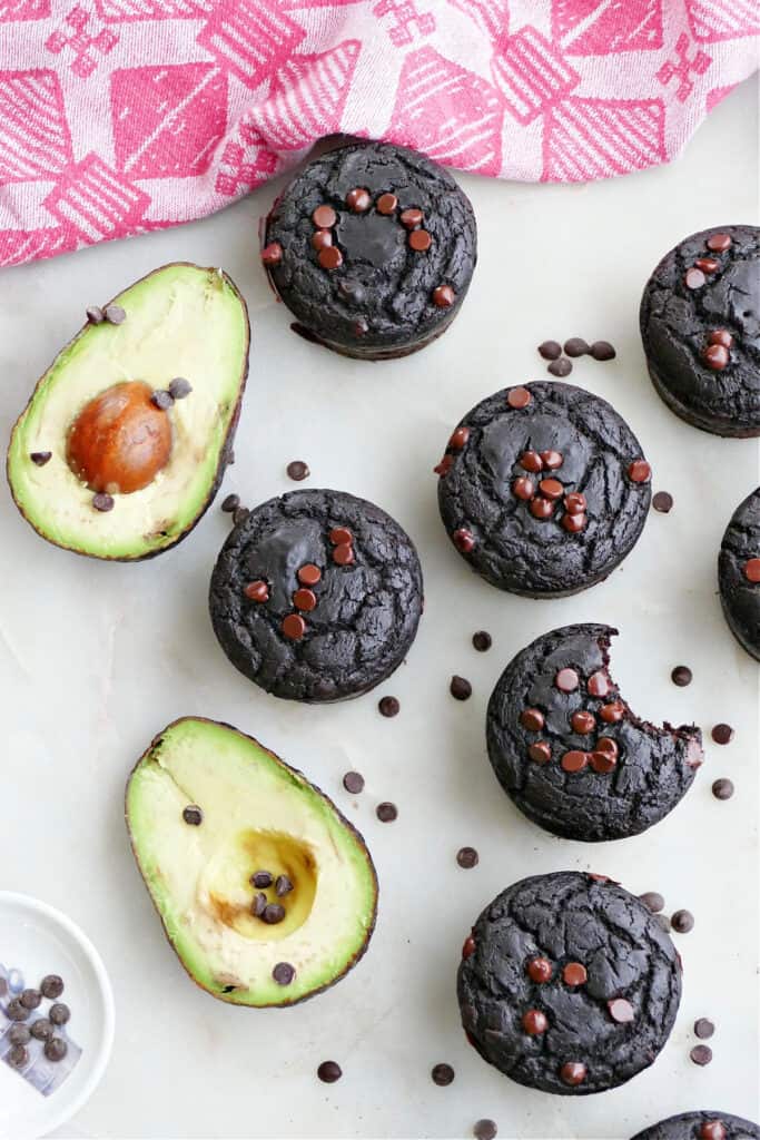 chocolate avocado muffins spread out next to avocados on a counter