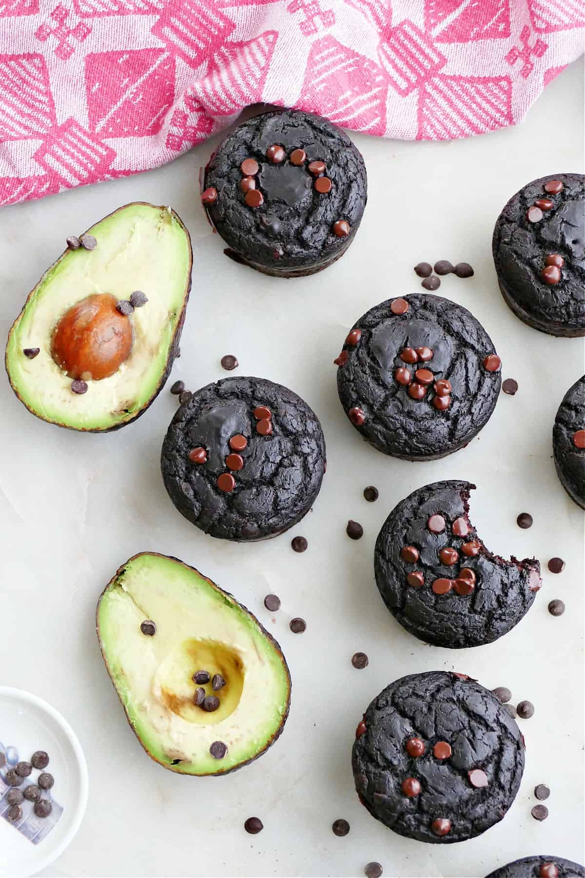chocolate avocado muffins spread out next to avocados and chocolate chips on a counter