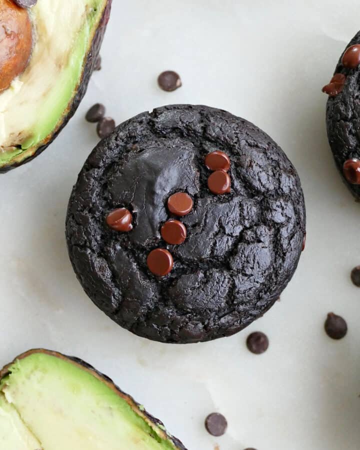 square image of a chocolate avocado muffin next to avocados on a counter