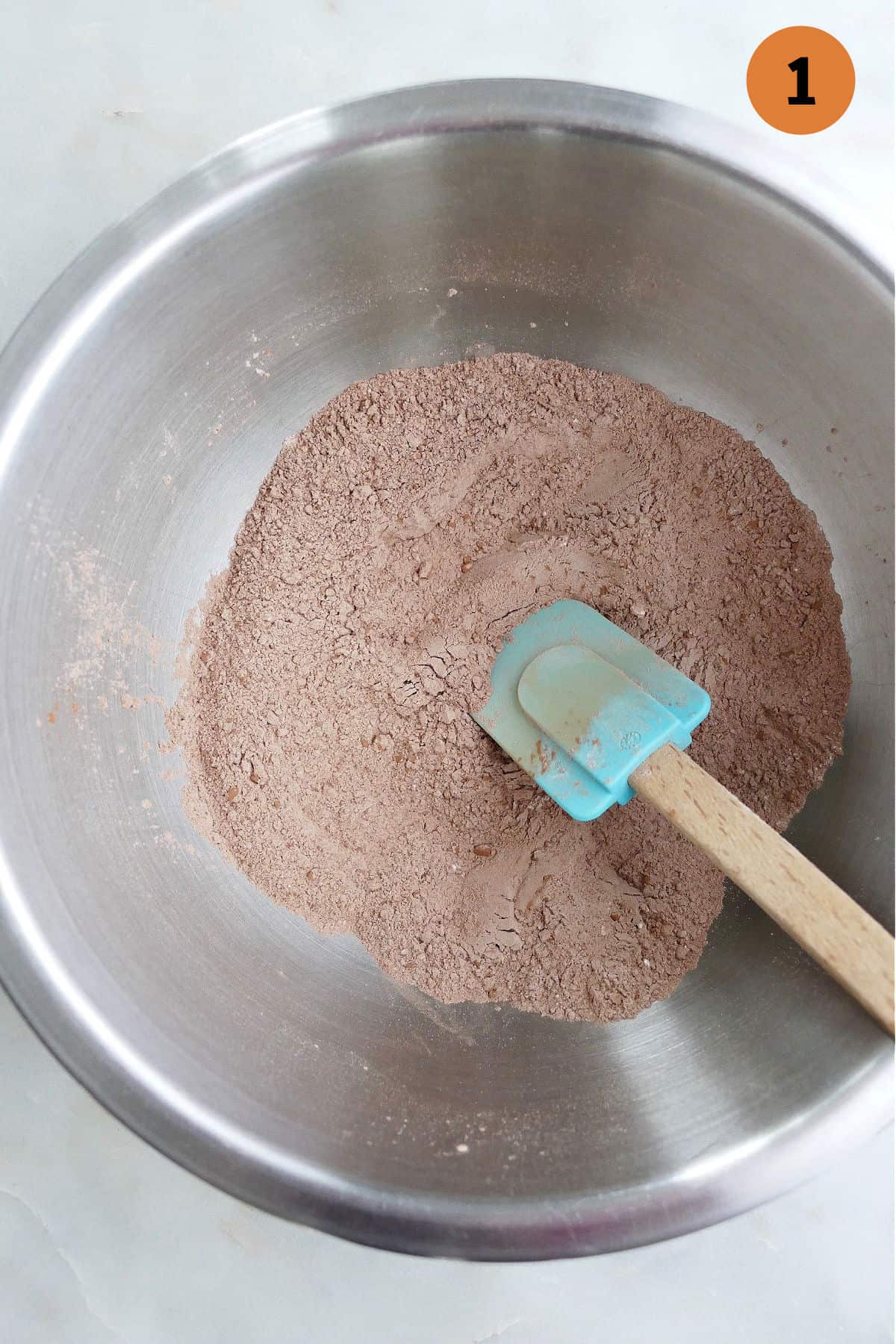 cocoa powder, flour, and other dry ingredients in a mixing bowl with a rubber spatula