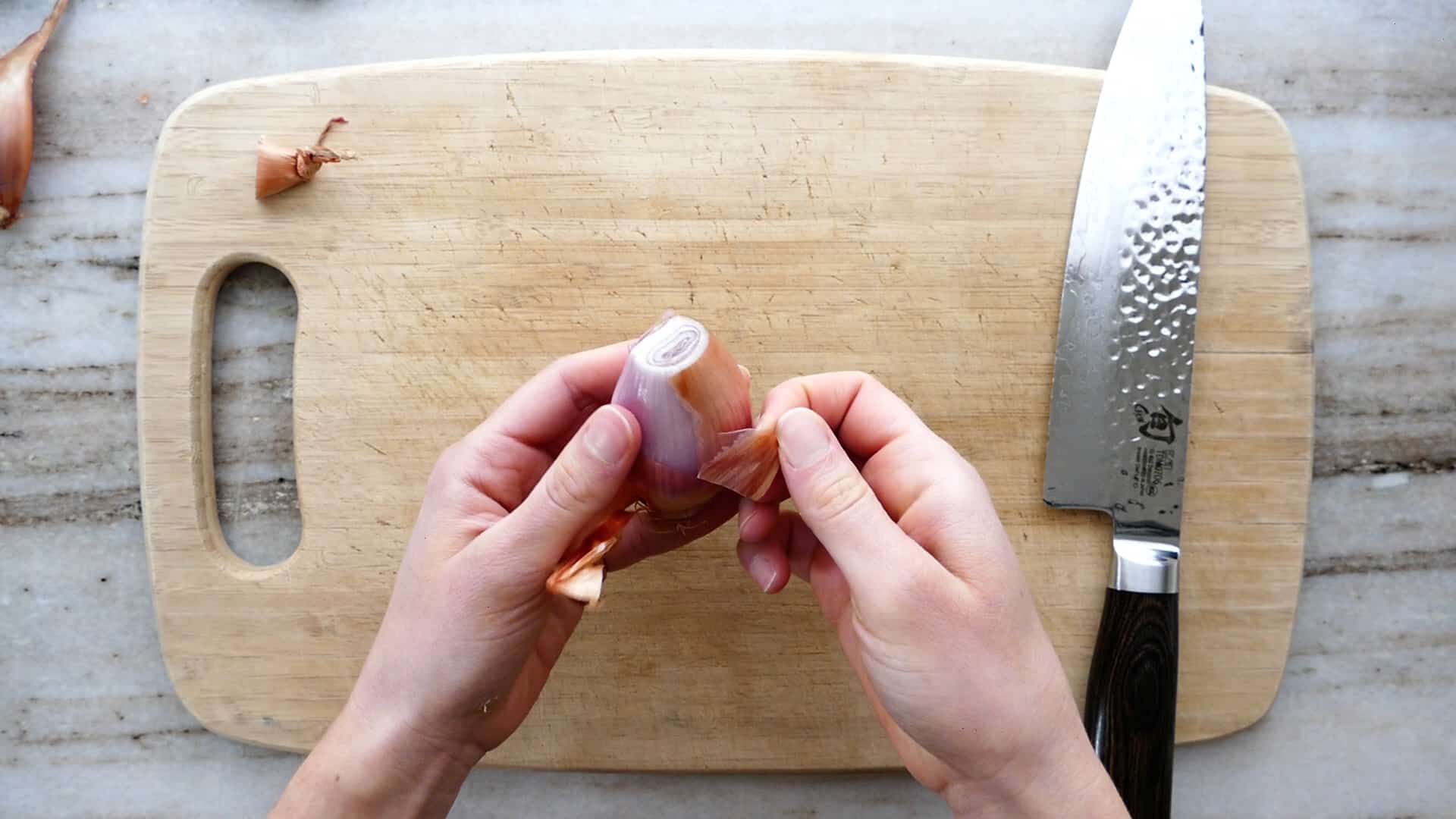 the hands of a woman peeling a shallot over a cutting board on a counter