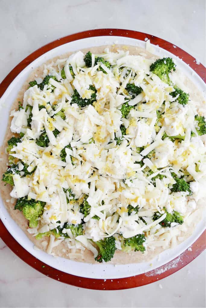 pizza dough on a pizza stone topped with cheeses, broccoli, and lemon zest