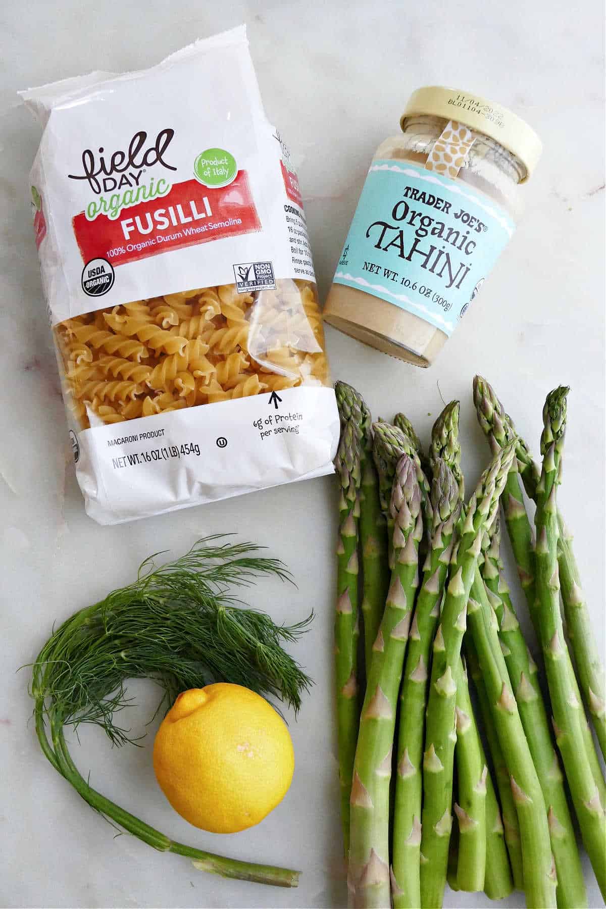 bag of fusilli pasta next to tahini jar, dill, lemon, and asparagus