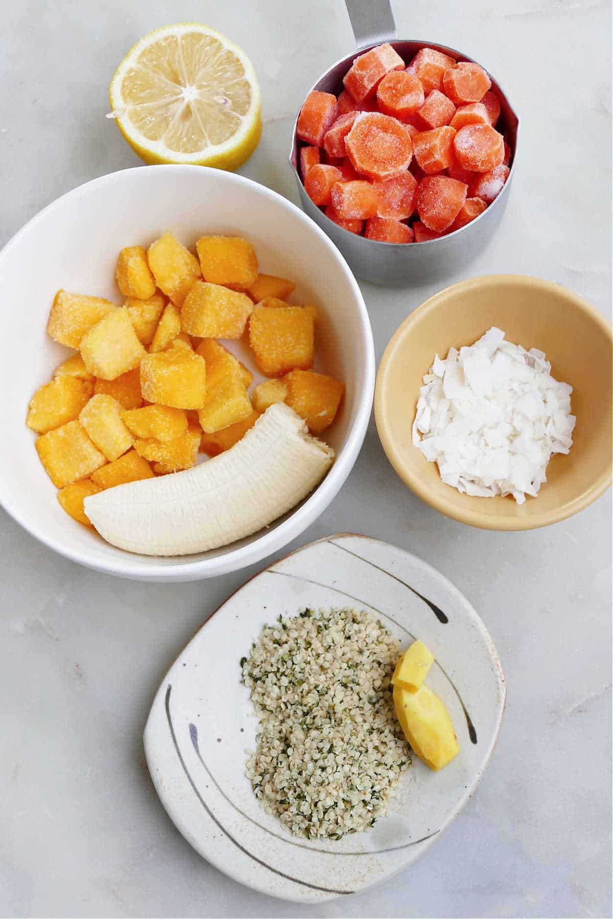 frozen carrots, mango, banana, hemp hearts, ginger, half a lemon, and coconut on a counter