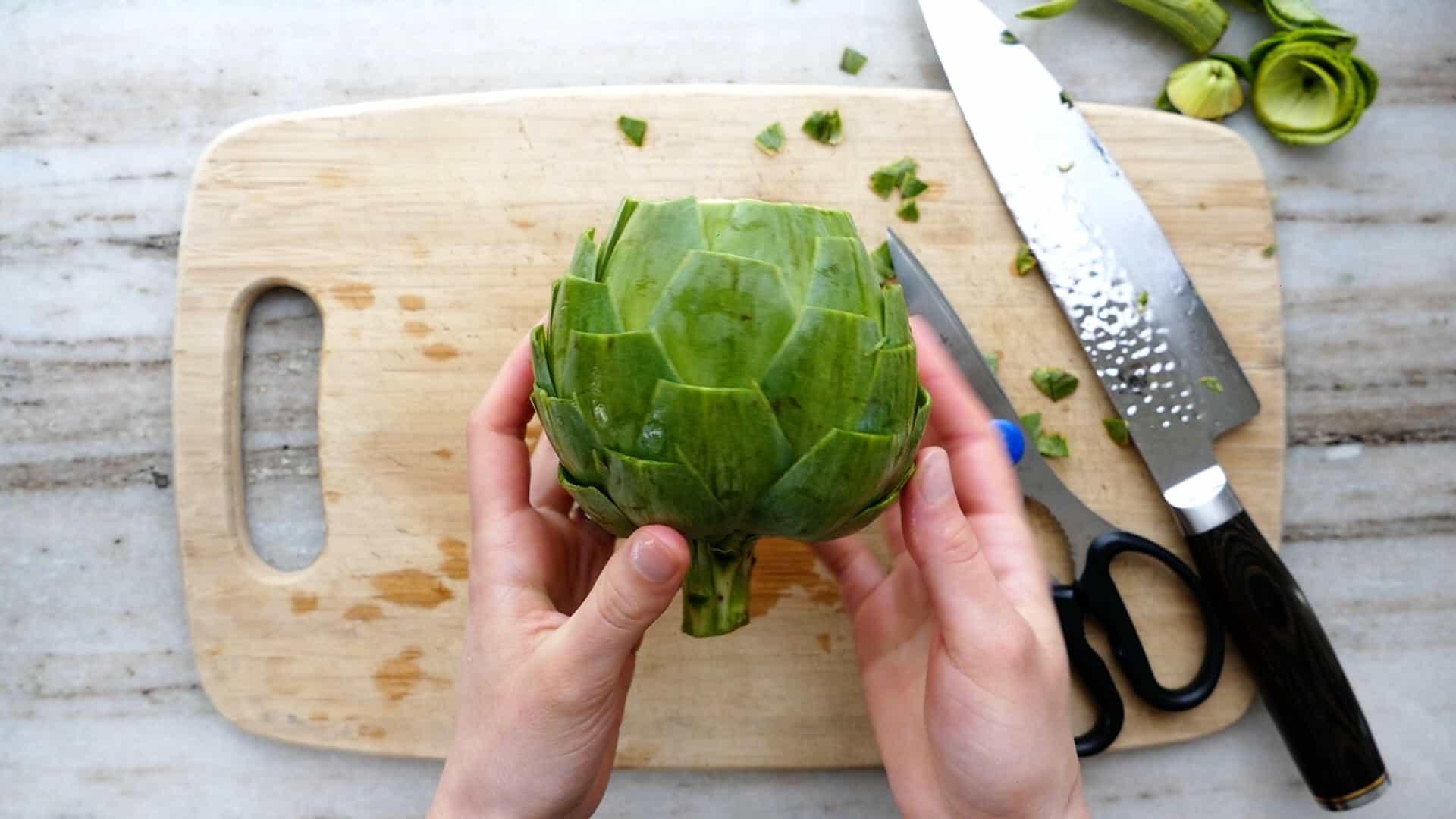 woman's hands holding up a trimmed artichoke over a cutting board
