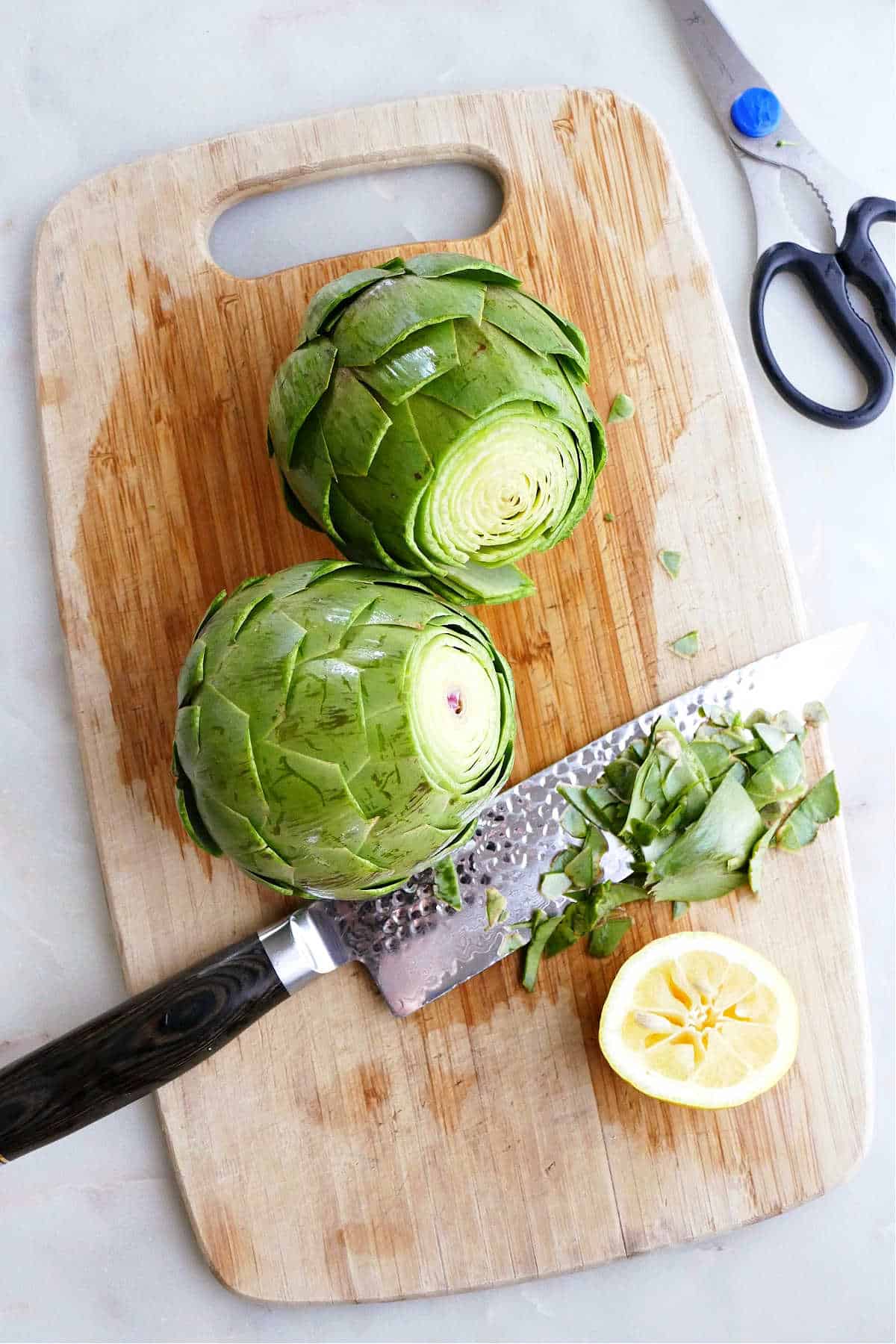 two artichokes trimmed with a knife on a cutting board next to a lemon