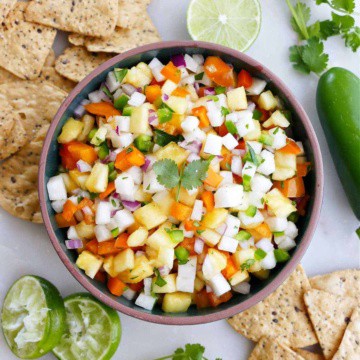 pineapple jicama salsa in a serving bowl surrounded by chips, lime, and cilantro