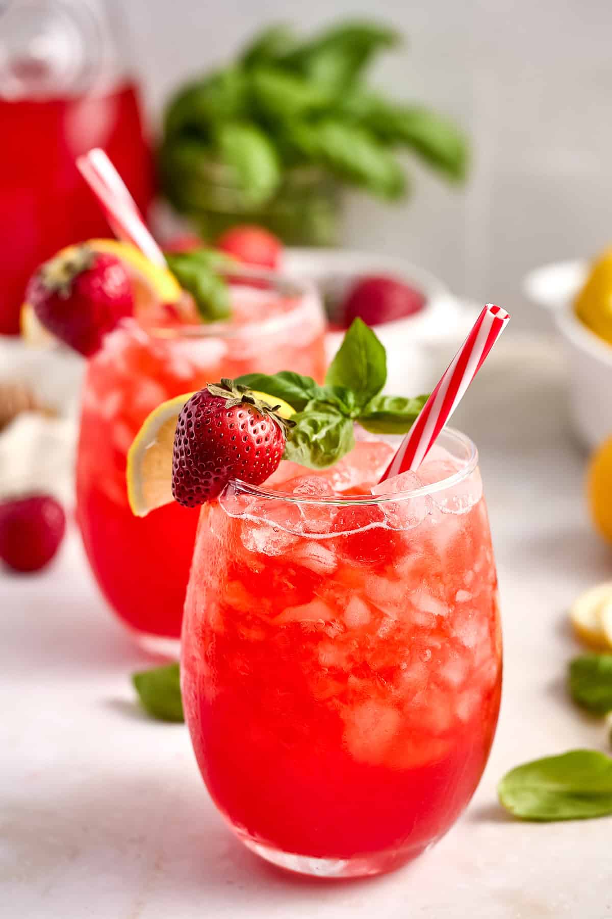Two glasses of strawberry lemonade with red and white straws and lemon, strawberry, and basil garnish.