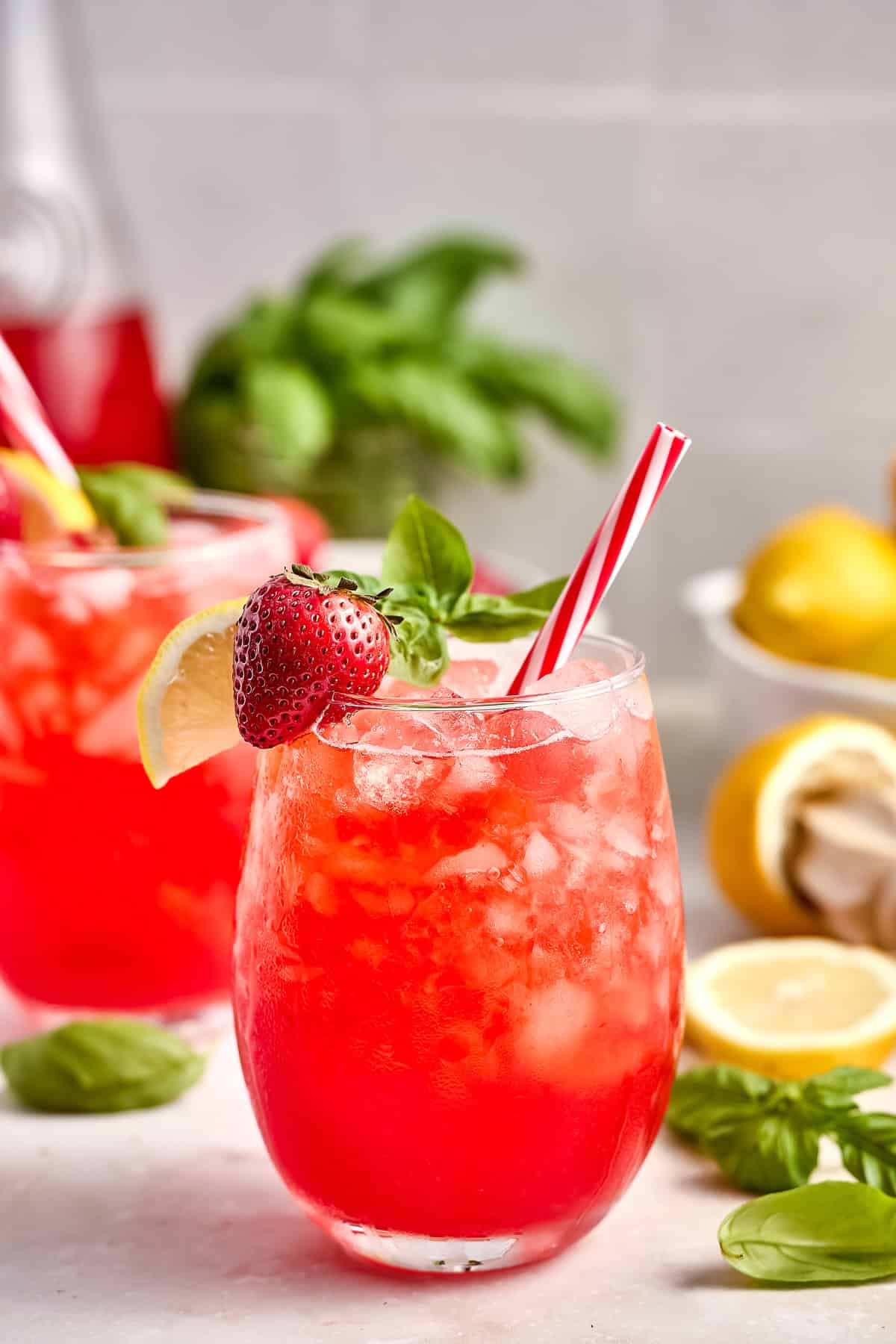 Strawberry basil lemonade in glasses with red and white straws surrounded by ingredients.
