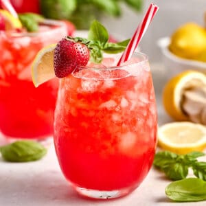Glass of strawberry basil lemonade with red and white straw and strawberry, basil, and lemon garnish.