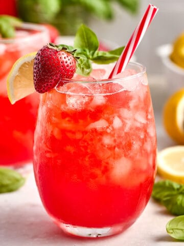 Glass of strawberry basil lemonade with red and white straw and strawberry, basil, and lemon garnish.