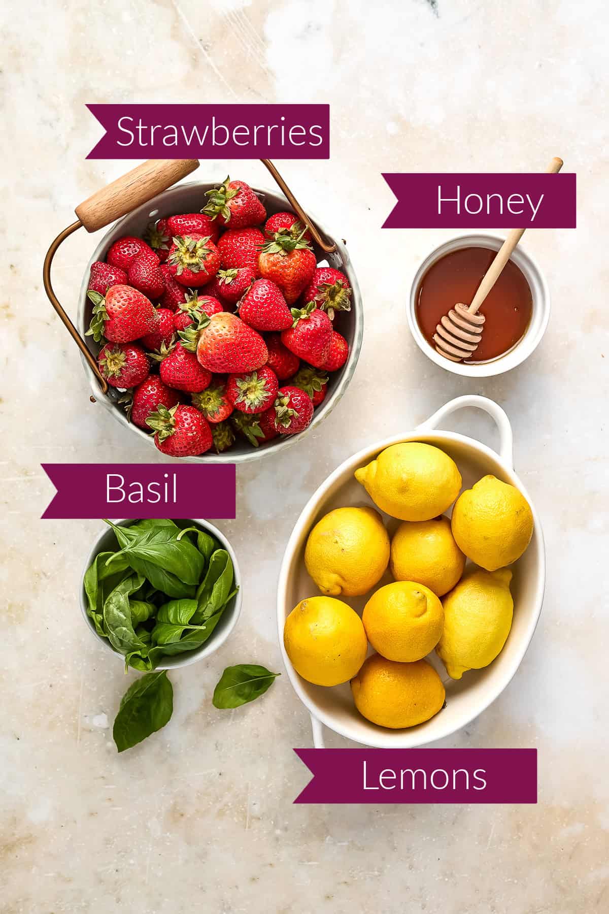 Strawberries, honey, basil, and lemons in bowls on a counter with labels.