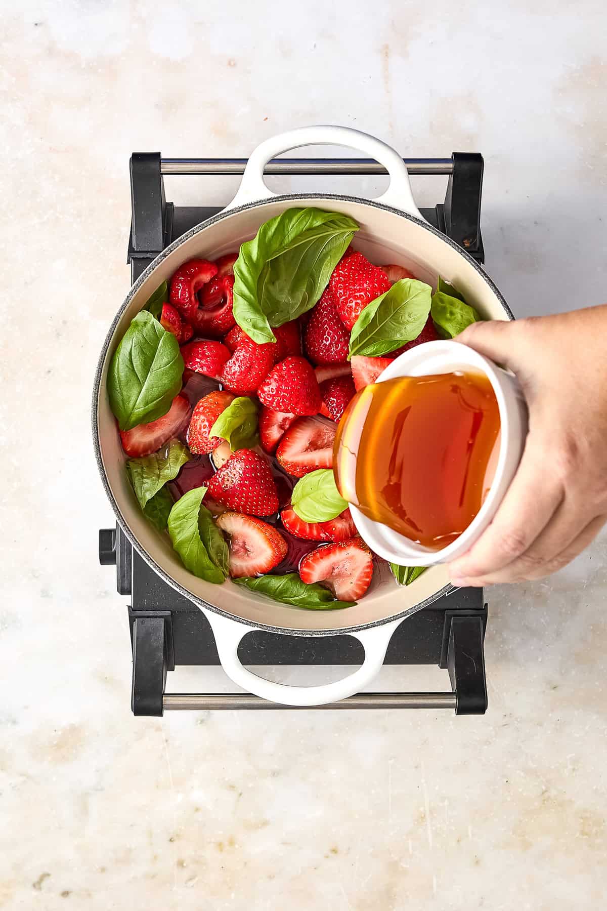 Woman pouring honey into a pot with strawberries and basil leaves over a stove.