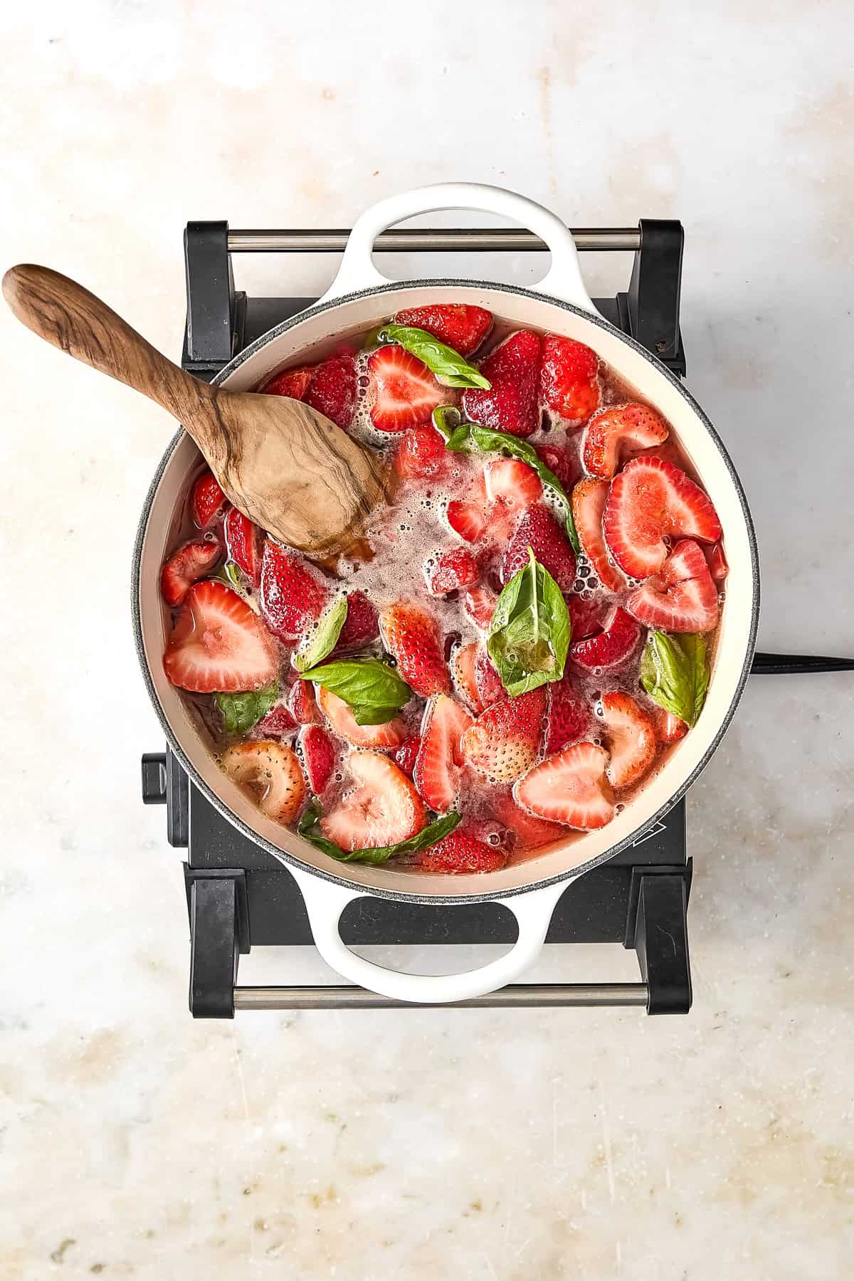 Strawberries, basil leaves, honey, and water in a large pot with a wooden spoon.