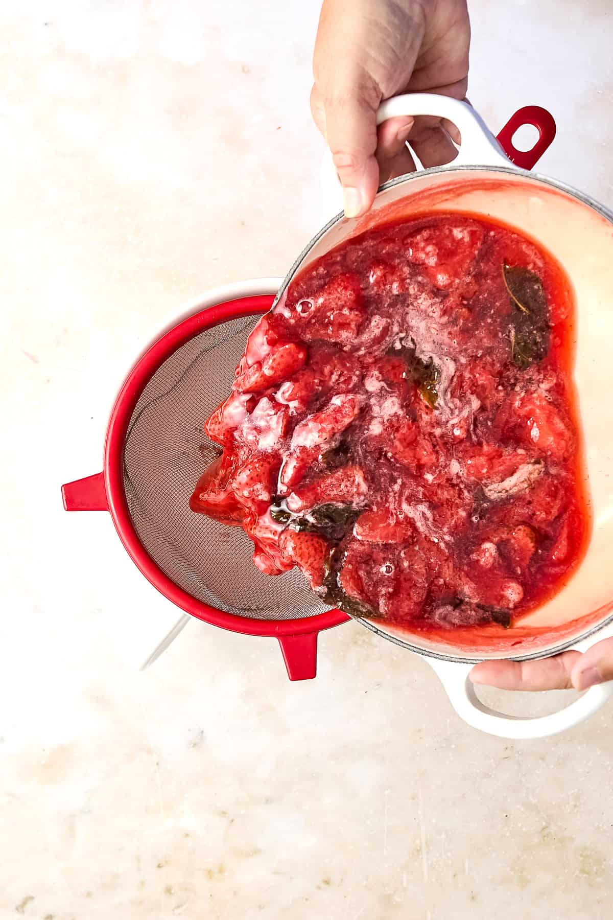 Woman pouring strawberry honey simple syrup for lemonade into a strainer over a bowl.