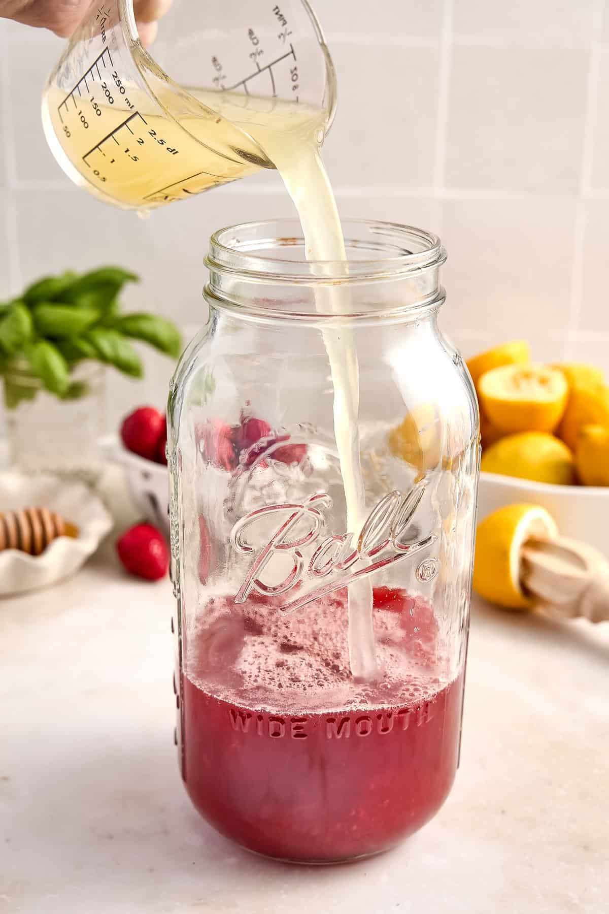 Woman pouring a measuring cup of lemon juice into a jar with strawberry syrup.