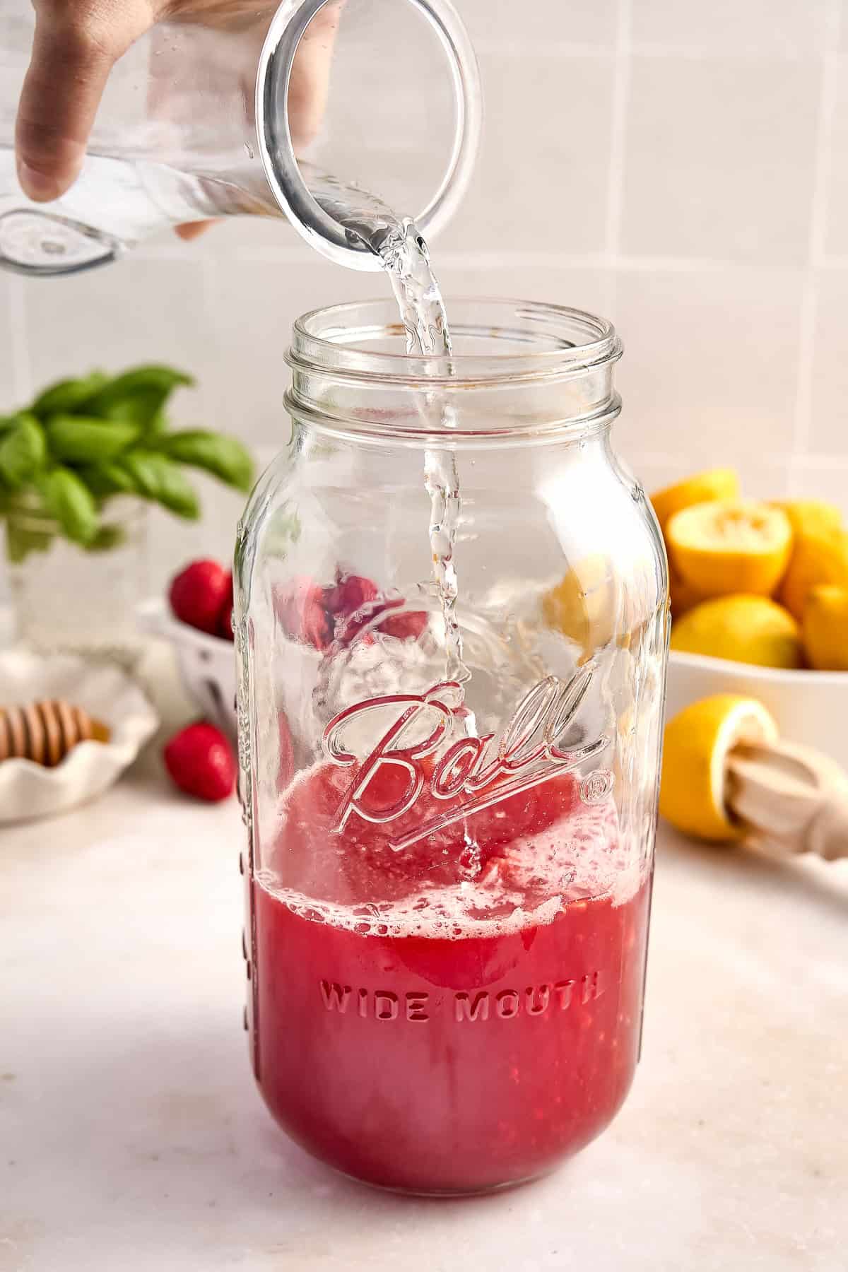 Woman pouring a measuring cup of water into a jar with strawberry syrup and lemon juice.