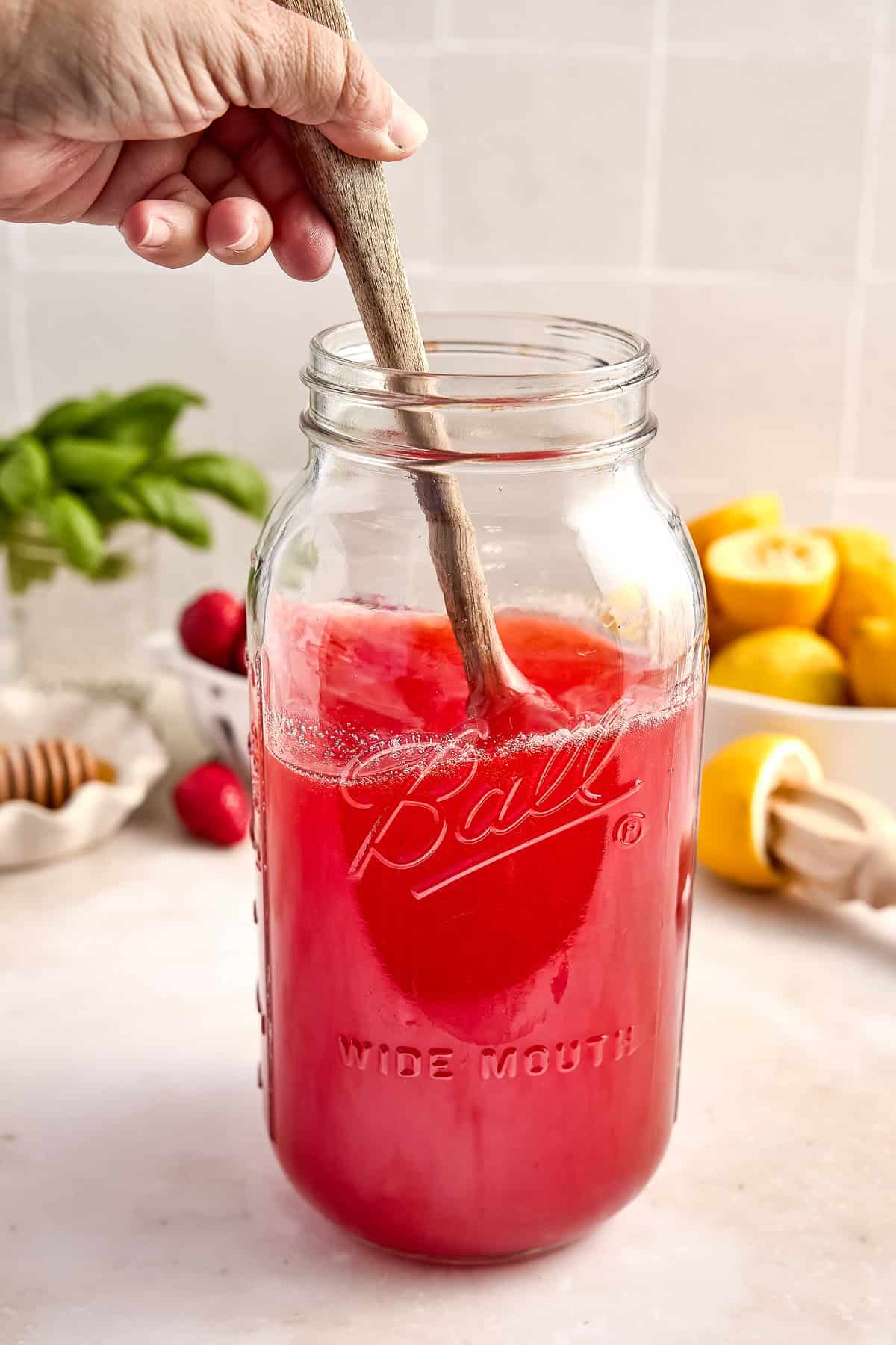 Woman stirring ingredients for strawberry basil lemonade in a jar with a wooden spoon.