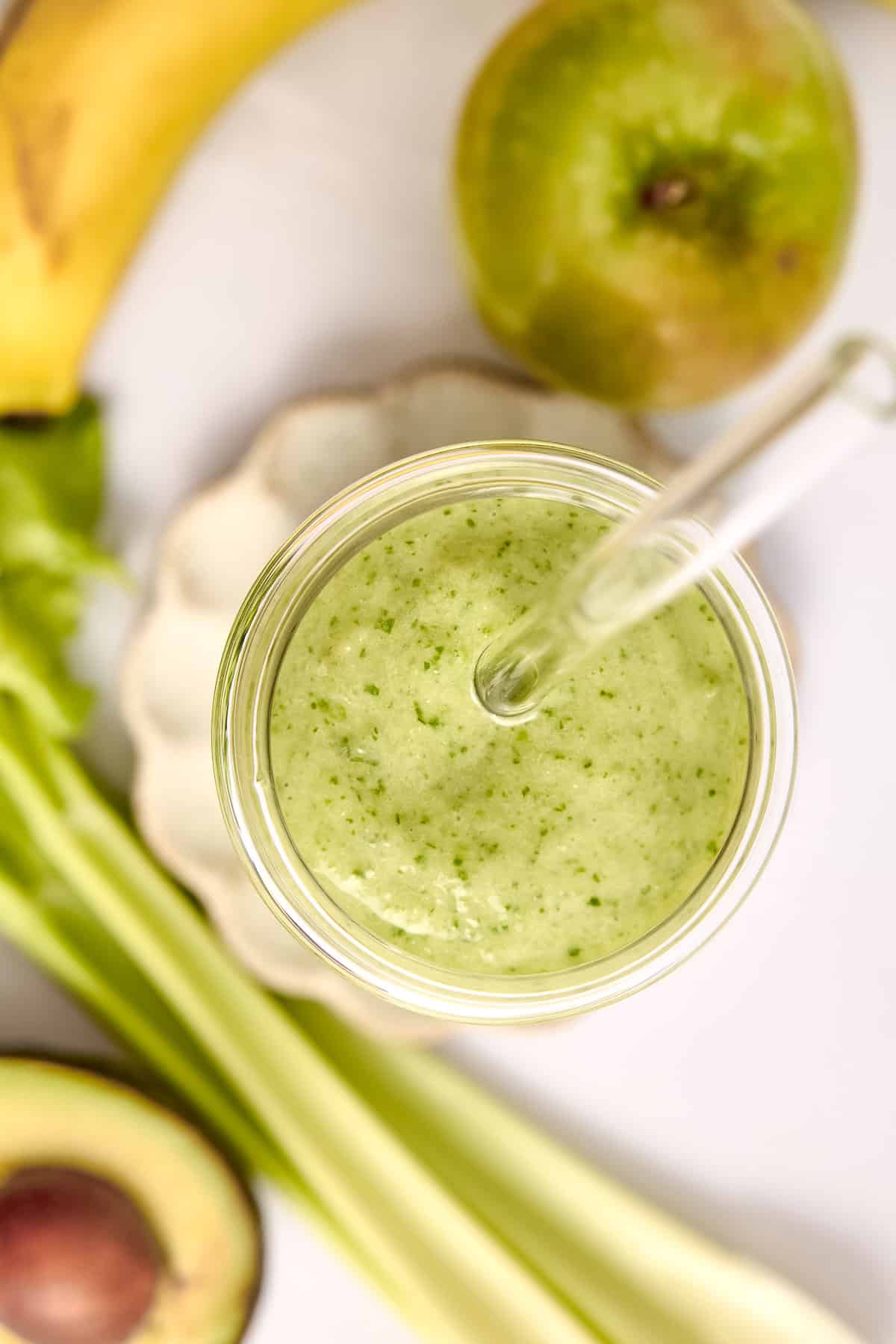 Top of celery smoothie in a glass with a straw next to ingredients.