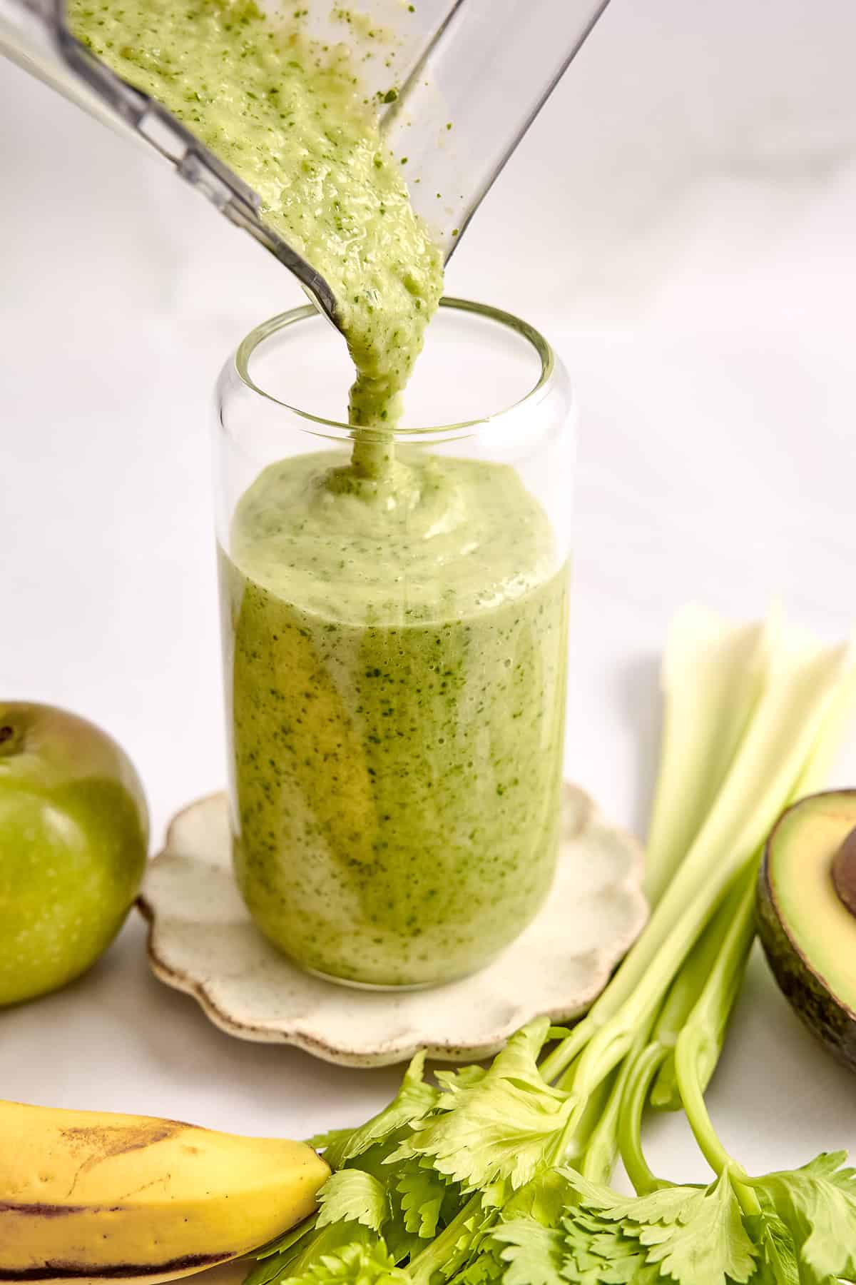 Celery smoothie being poured into a glass from a blender surrounded by ingredients.