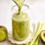 Celery smoothie being poured into a glass surrounded by ingredients.
