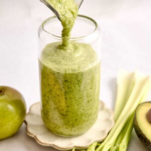 Celery smoothie being poured into a glass surrounded by ingredients.